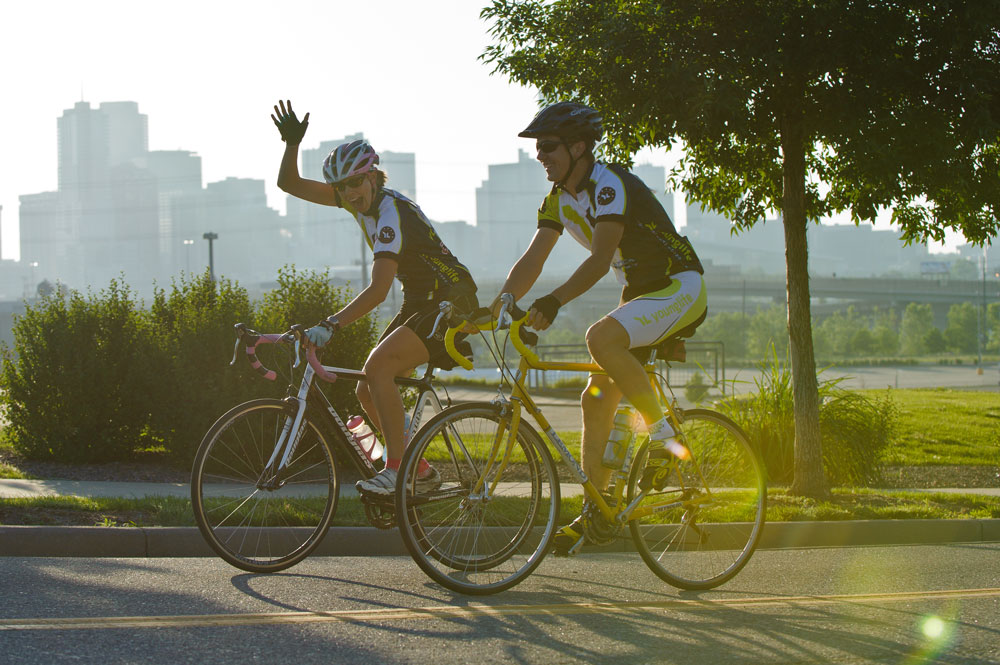 Two people riding on a road toward the left side of the frame. One is waving and smiling. There is a city skyline in the background.