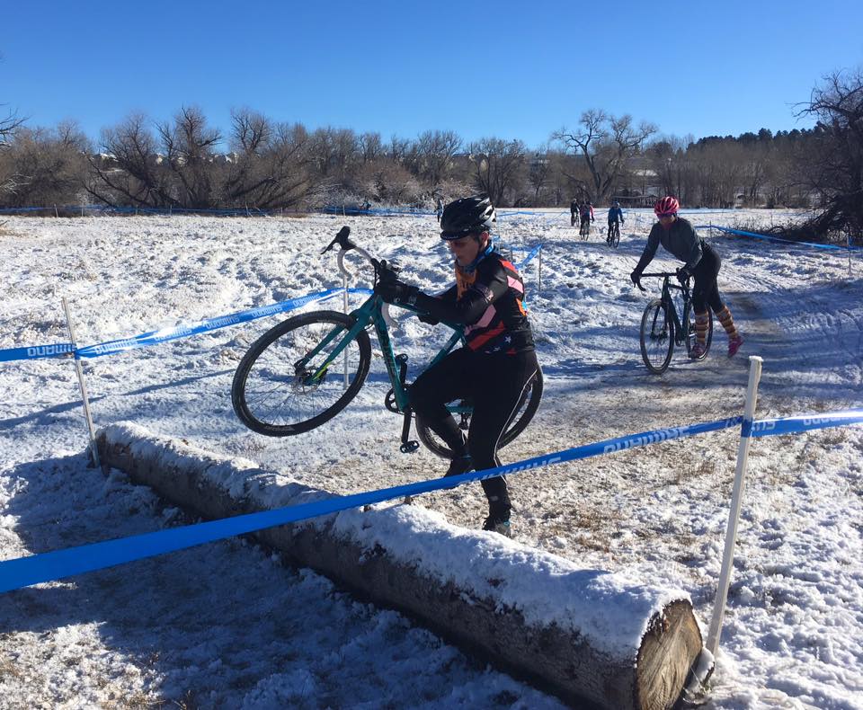 A person lifting their bike over a snow covered log on dirty and snow covered ground in a cyclocross race.