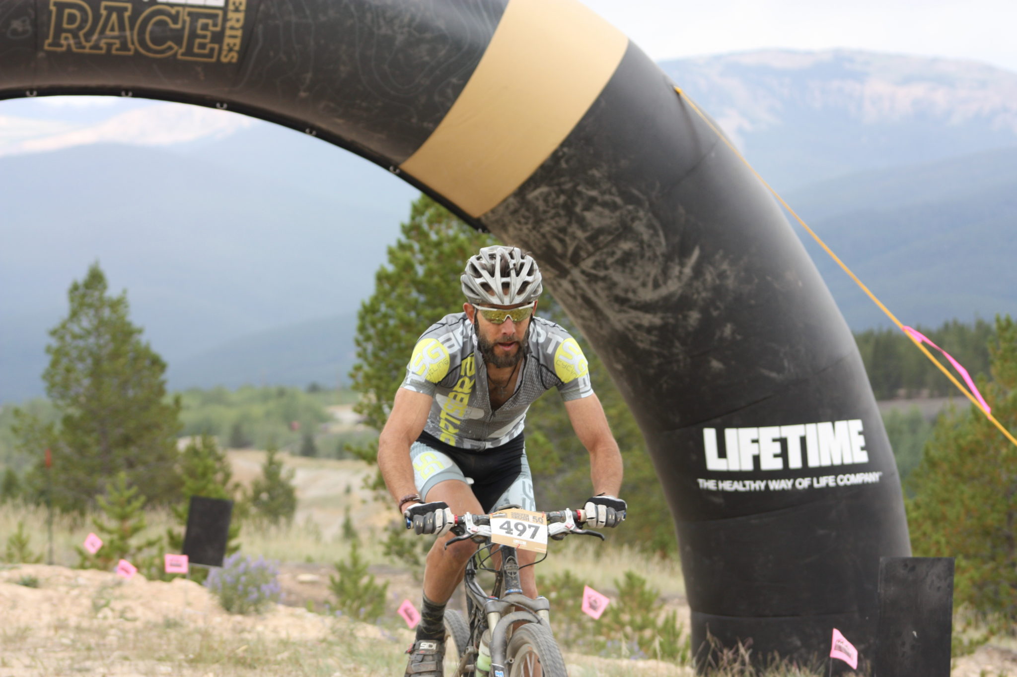 A white man riding a mountain bike passes through a finish line arch with tall hills and mountains in the unfocused background