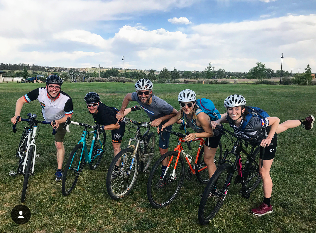 Five people, Bicycle Colorado team members, stand with their bikes, each of them with their right legs up and back behind them, on a grass field.