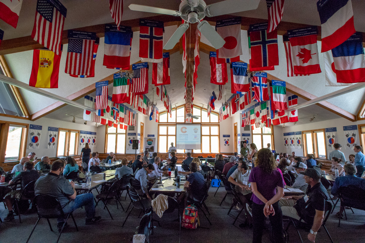 View from the back of a large community room with national flags hanging from a high ceiling. There are many people seated at long tables looking toward the front of the room, away from the camera, where a person is speaking at a podium and projecting a presentation on a screen behind them.