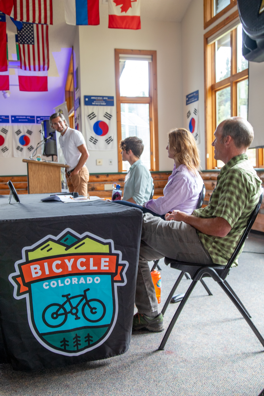 Three people sitting at a table with a cloth with the Bicycle Colorado logo on it looking over at a man speaking at a podium. They are indoors in a high ceilinged space with flags hanging from the ceiling.