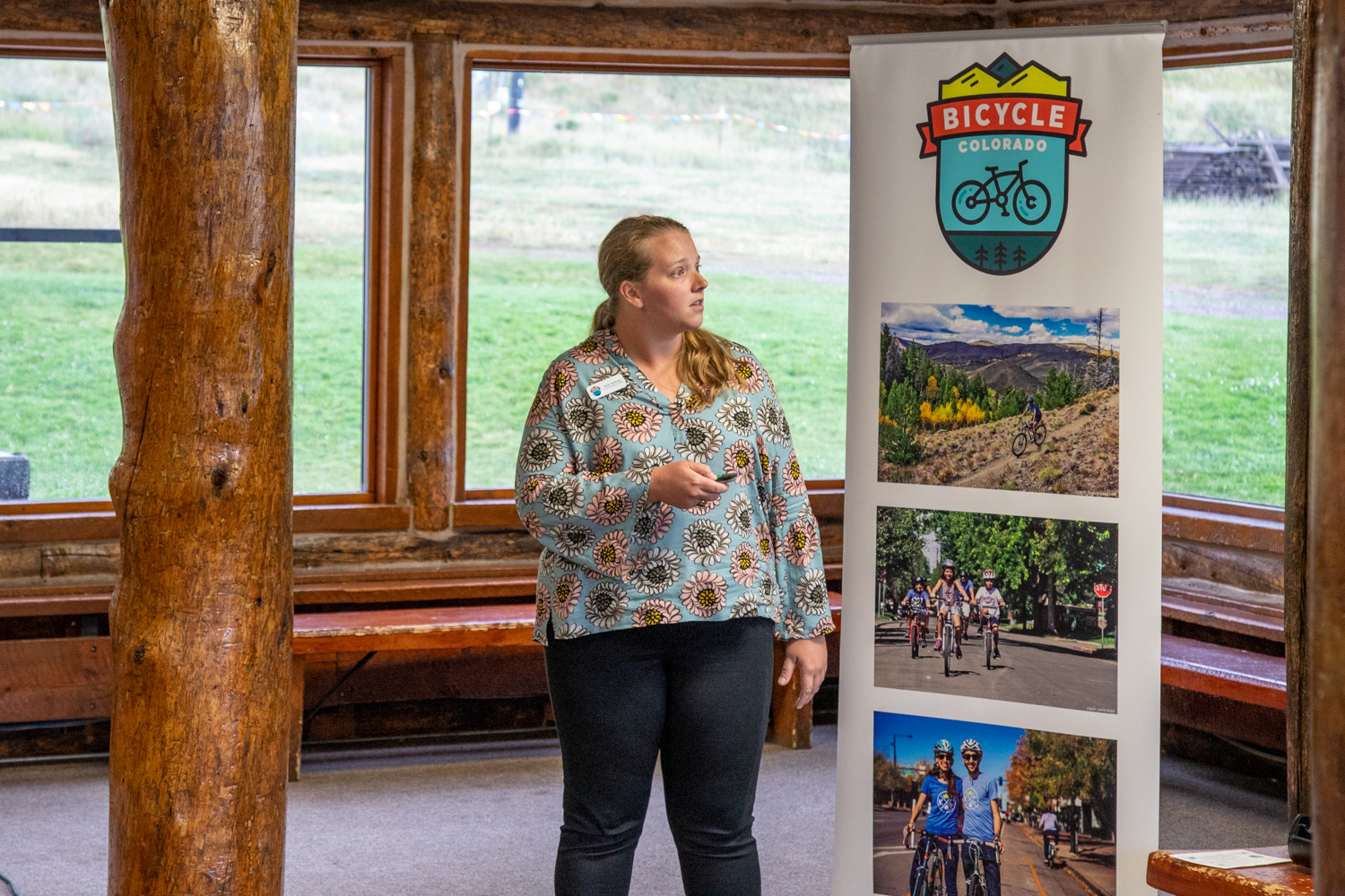 A white woman looks to the right to a presentation offscreen, standing next to a vertical banner with photos of people bicycling and the Bicycle Colorado logo.