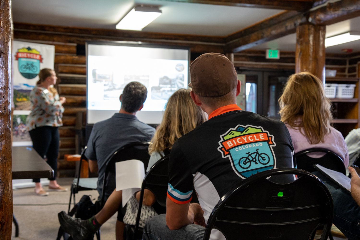 A person wearing a Bicycle Colorado jersey sitting in a chair in the audience of a presentation in the background.