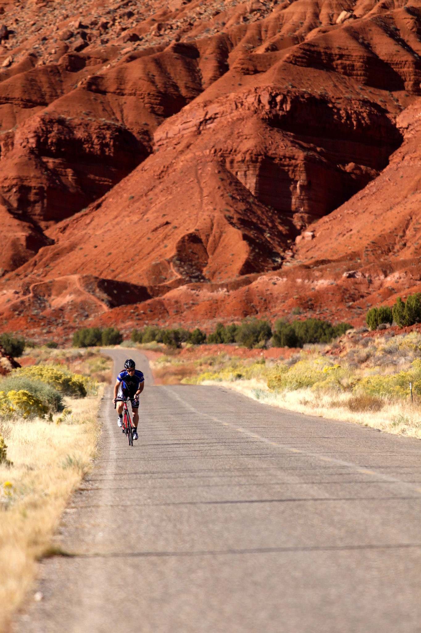 A person riding a bike on a paved road toward the camera with red rock hillsides looming in the background