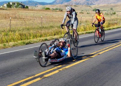People riding bikes on a paved road through farmland. The person in the front is riding a fully recumbent hand cycle and the other two are riding traditional upright bikes.