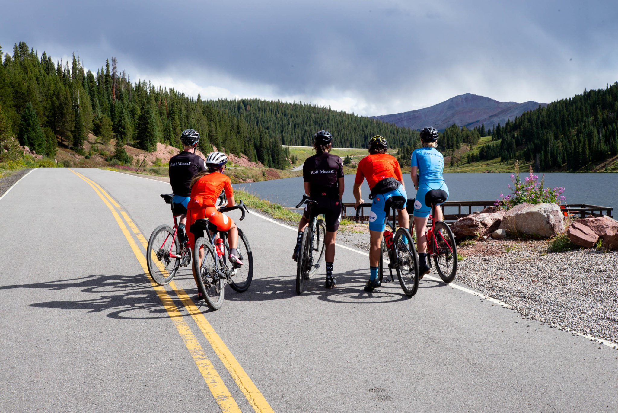 Five people stopped standing over their bikes on a paved road looking at a lake and a mountain.