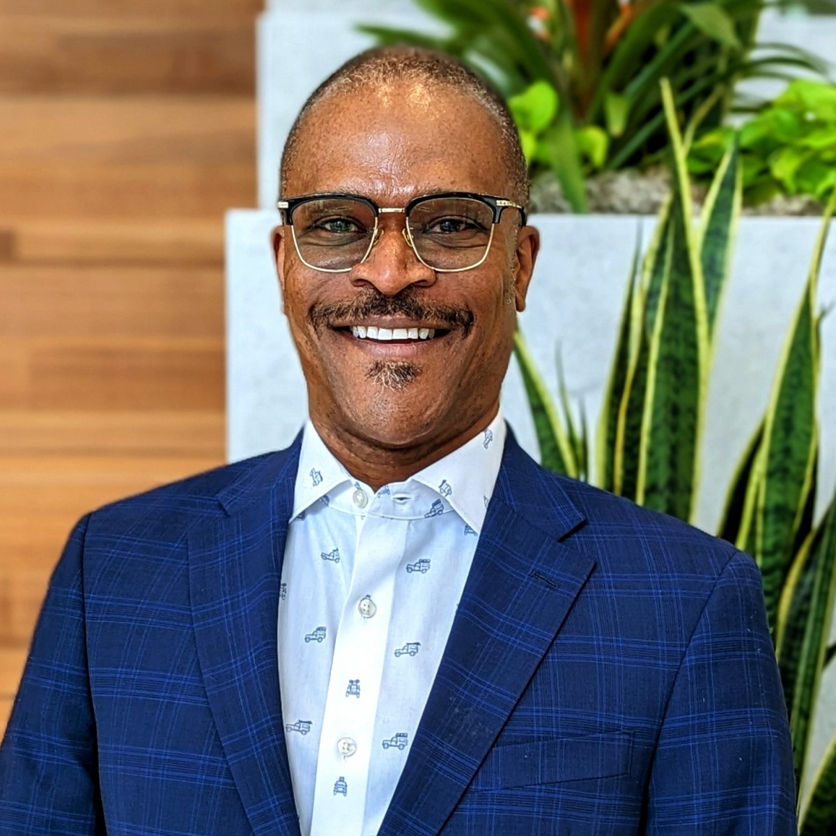 A Black man with a moustache and very short hair, photographed from the chest up in a blue suit jacket and white shirt.