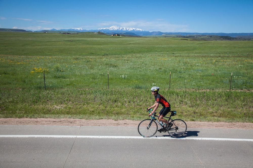 A person riding a bike on a road shoulder toward the left of the frame, next to a grassy field.