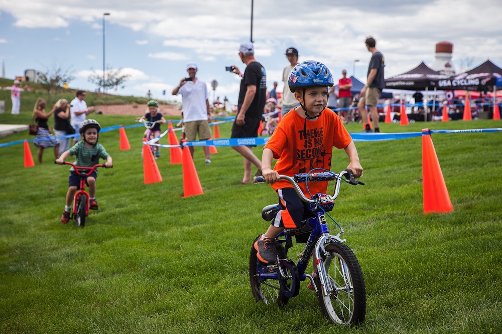 Children riding bikes on grass on a course marked by cones and plastic tape while adults look on.