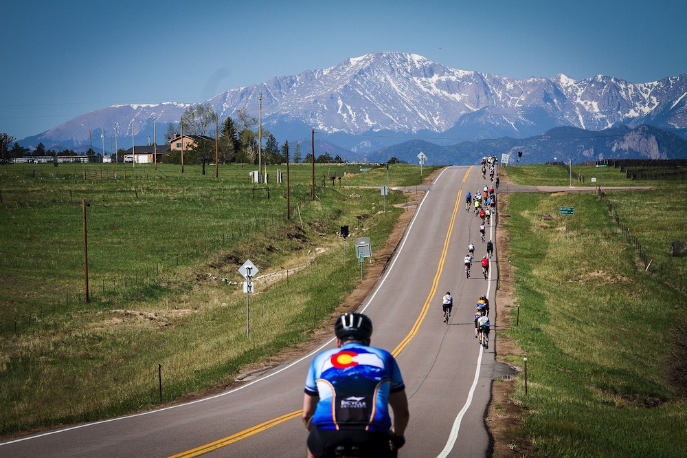 People riding bikes on a paved road away from the camera with large mountains in the background.