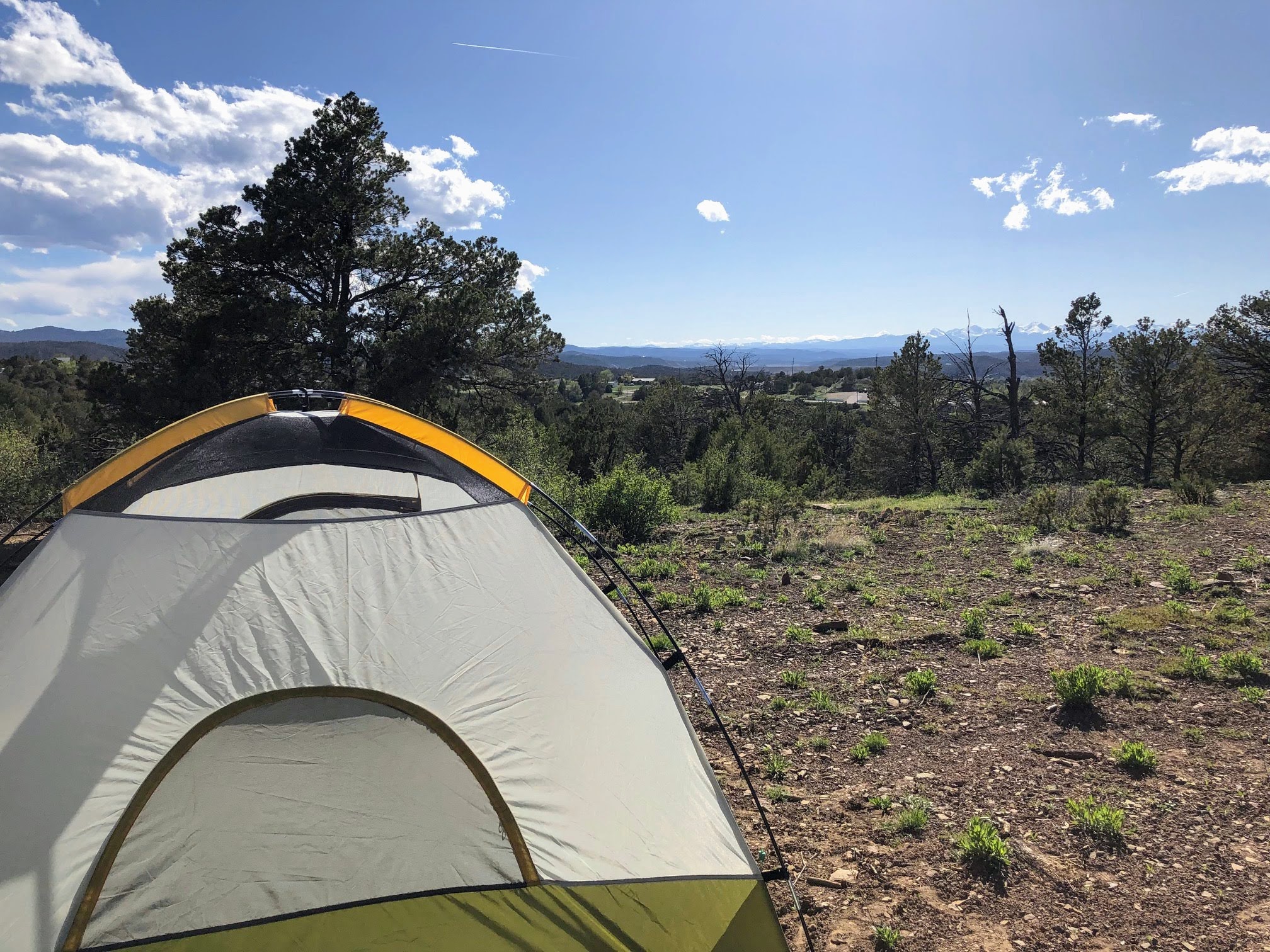 A tent pitched on a dirt lot with trees and large mountains in the far background.