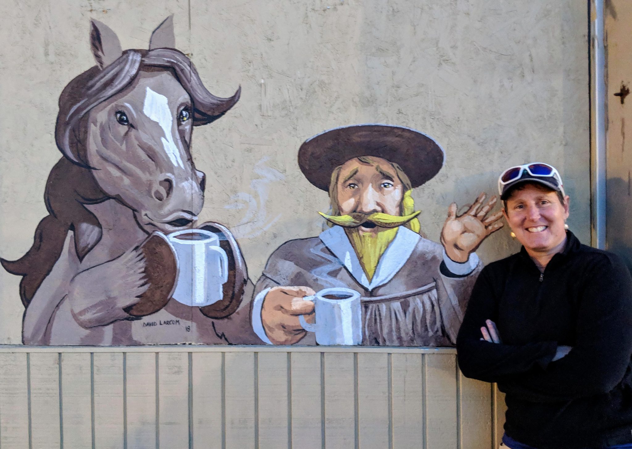 A person posing with a mural depicting a horse and a human both holding cups of coffee.