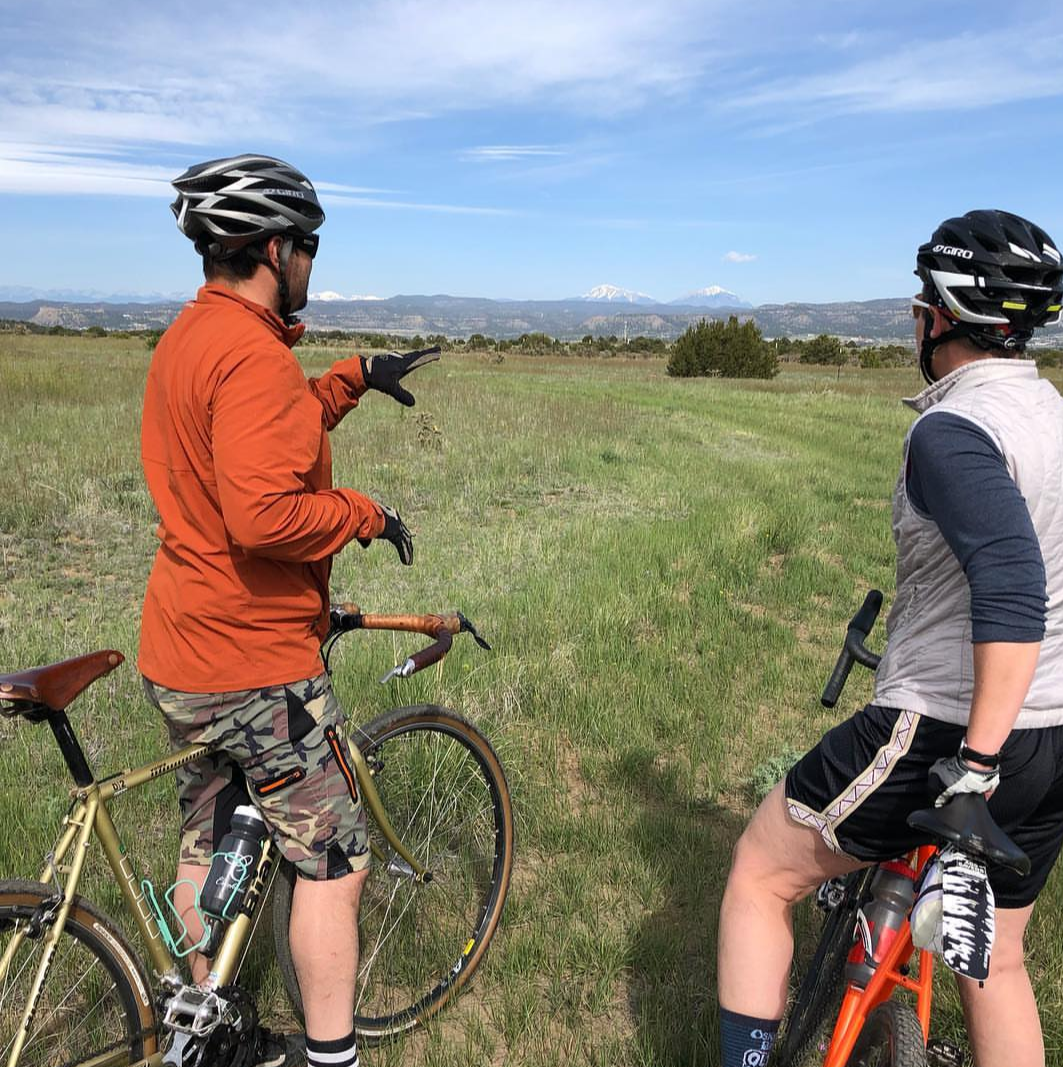 Two people standing over bikes look over a field at large mountains in the background.