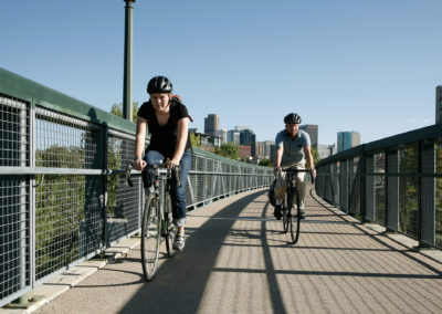 People bicycling on a bike and pedestrian bridge with the Denver skyline behind them.