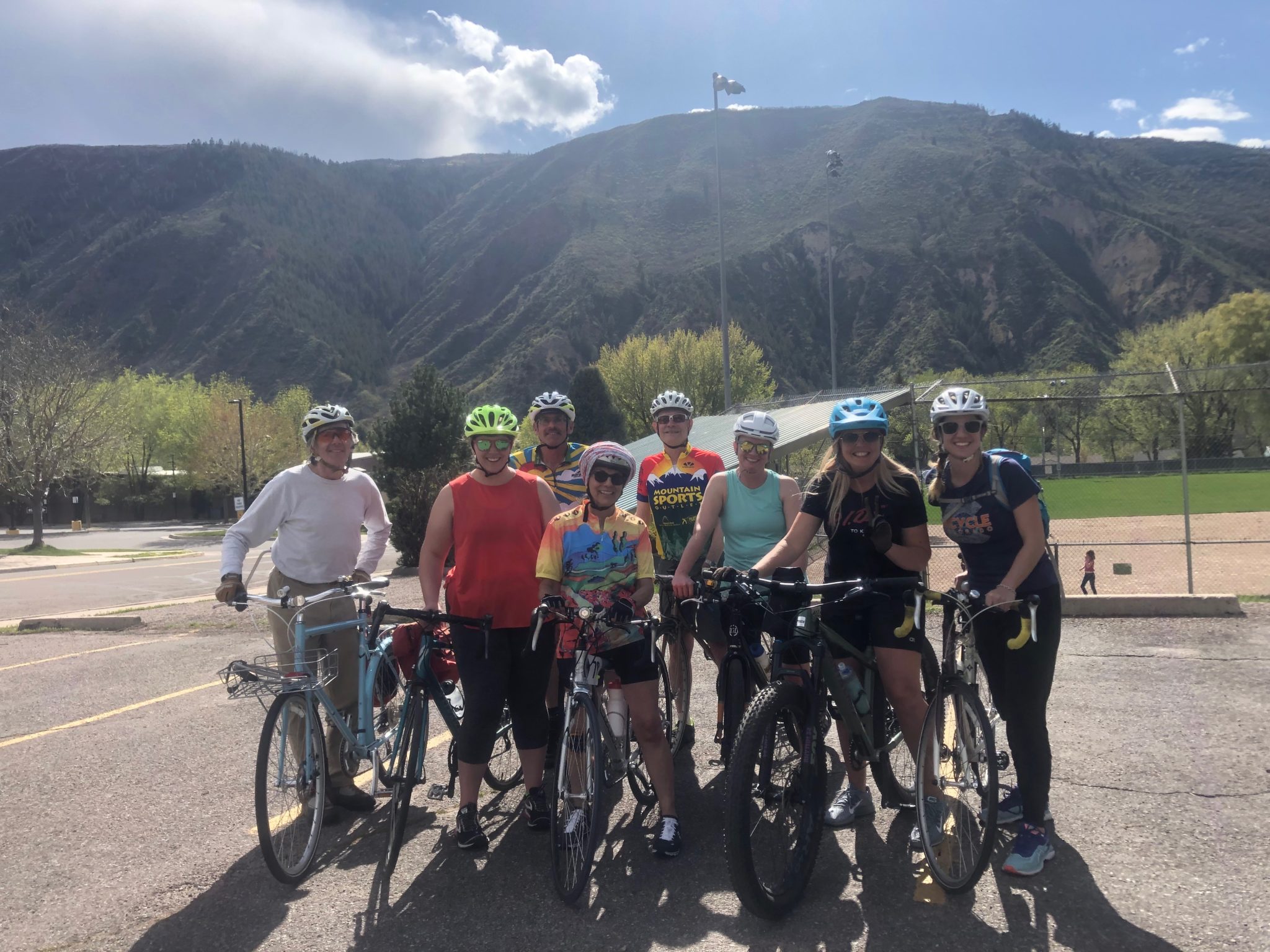 Eight people pose with bicycles in a parking lot below large hills.