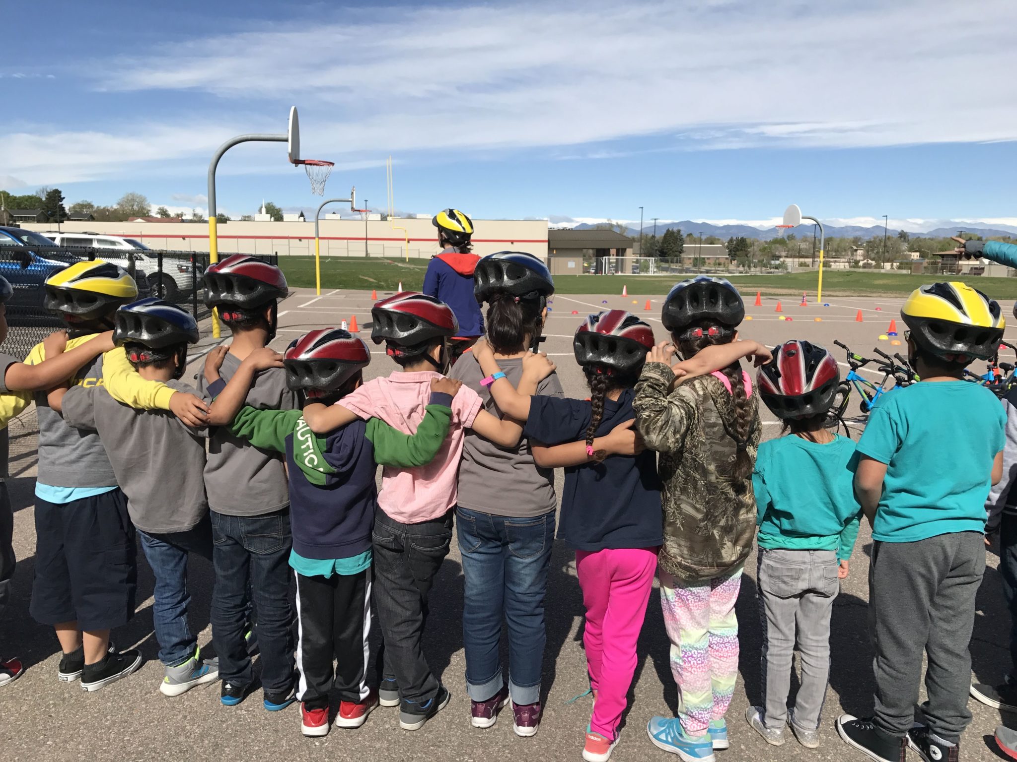 A group of children stand with their arms around each other and backs to the camera. They are wearing helmets and are in a school playground.