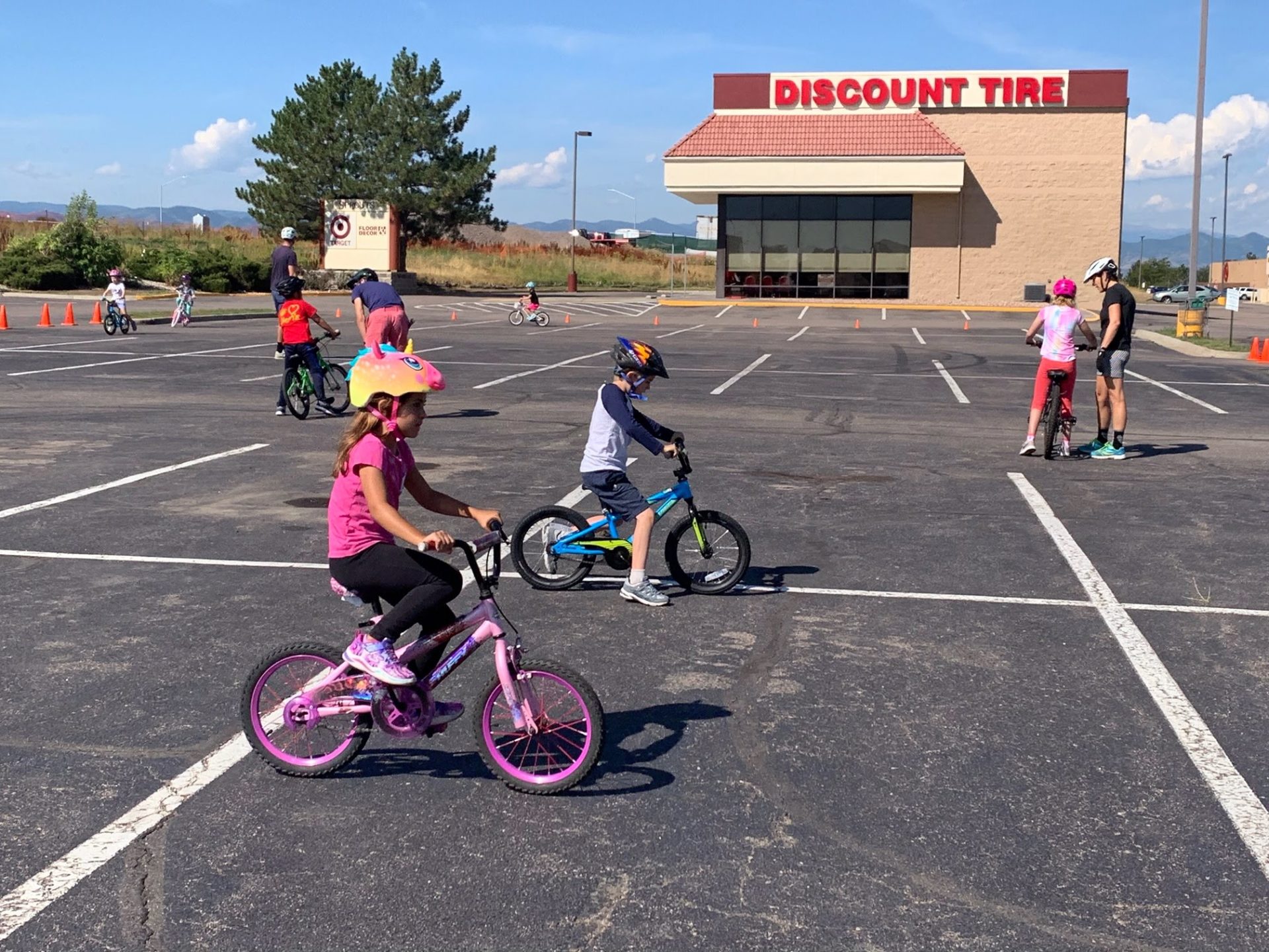 A number of children learning to ride bikes, with adults nearby instructing them, in a parking lot.