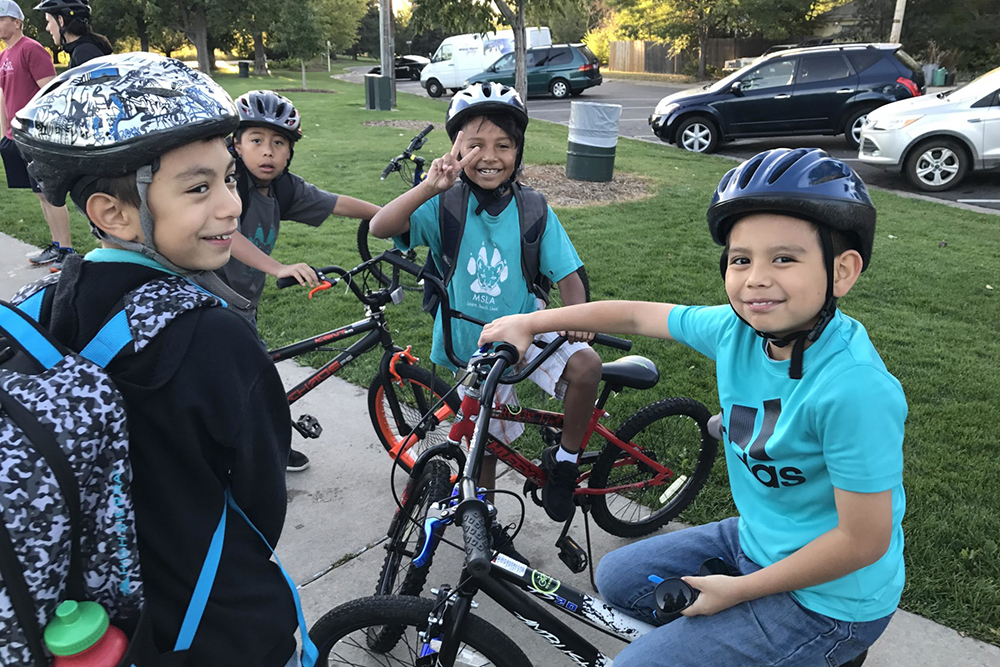 Four children stand facing each other but turned toward the camera. They are wearing bike helmets and a couple of them have bikes. They are on a sidewalk.