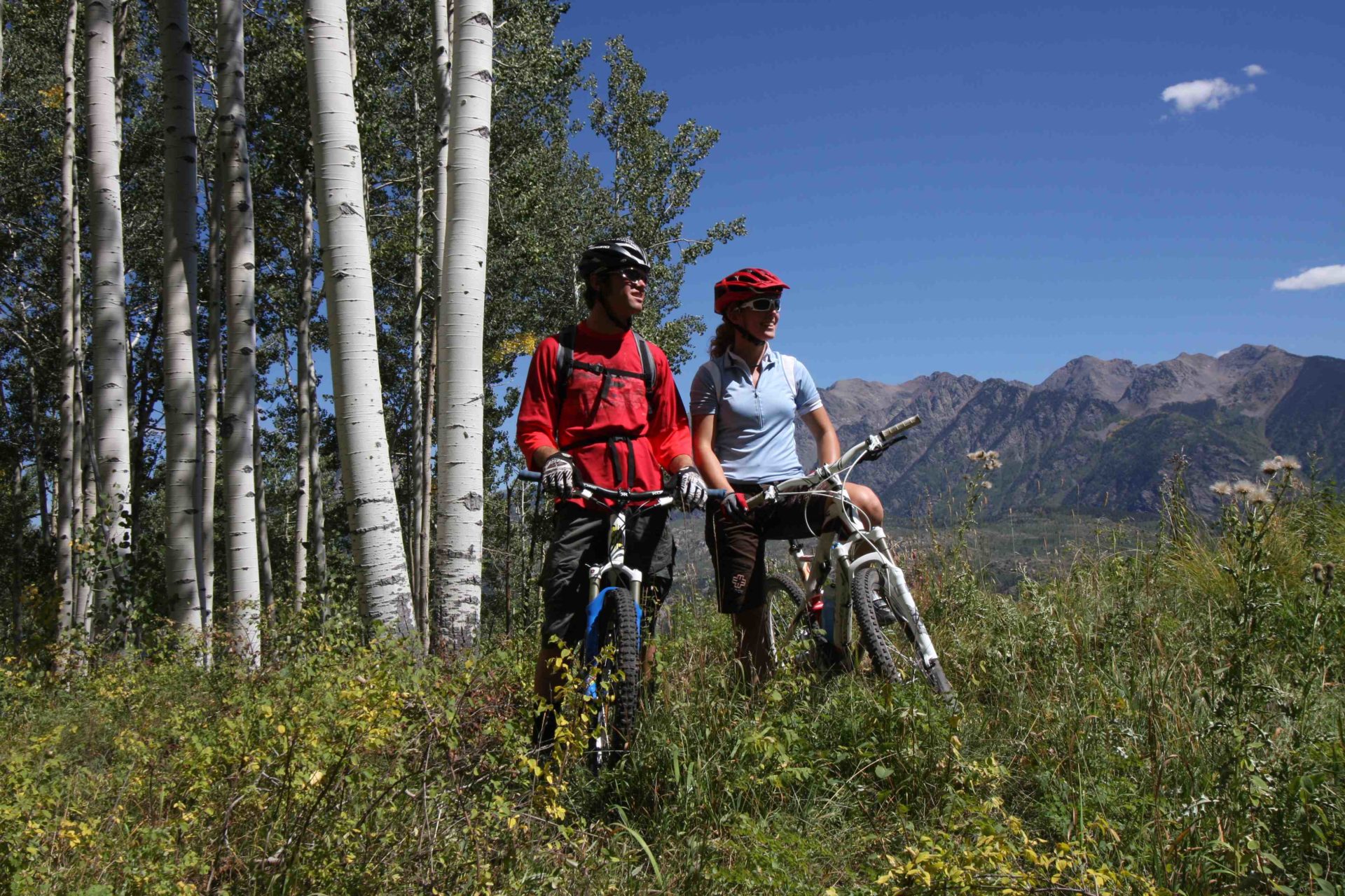 Two people stand and smile astride their bikes next to an aspen grove with mountains in the background.