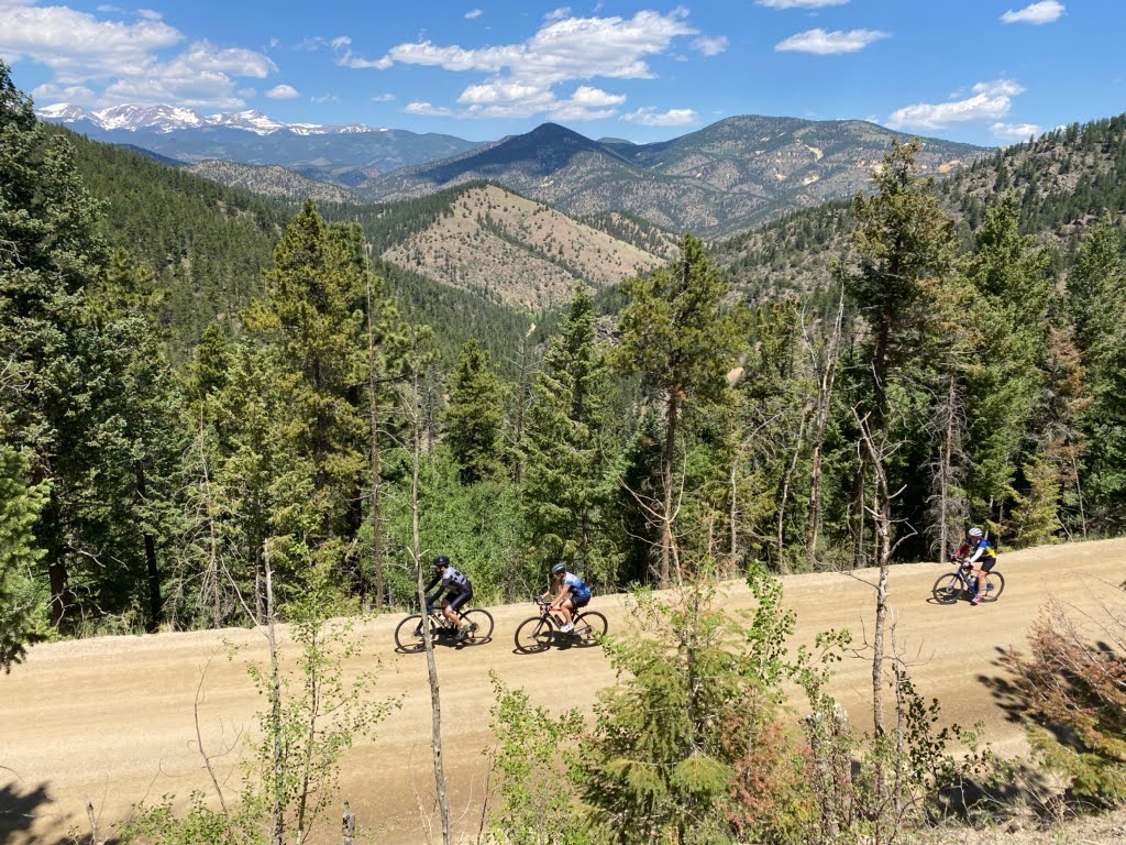 People riding a bike on a dirt trail, with a view of mountains behind them. They are heading to the left.