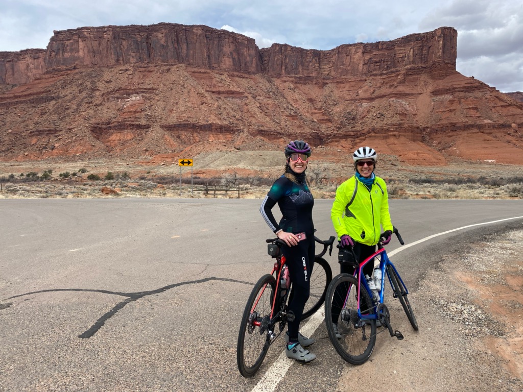 Mary and Nadine pose with their bikes on a road in front of red rocks in Moab.