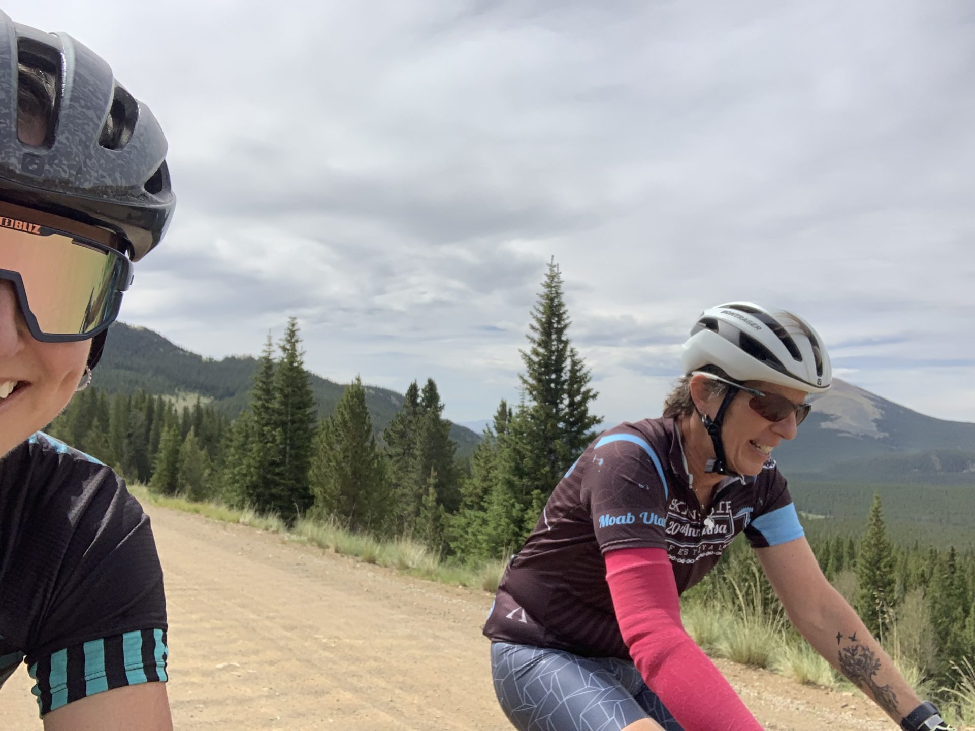 Mary and Nadine wearing riding kits and helmets ride bikes on gravel.