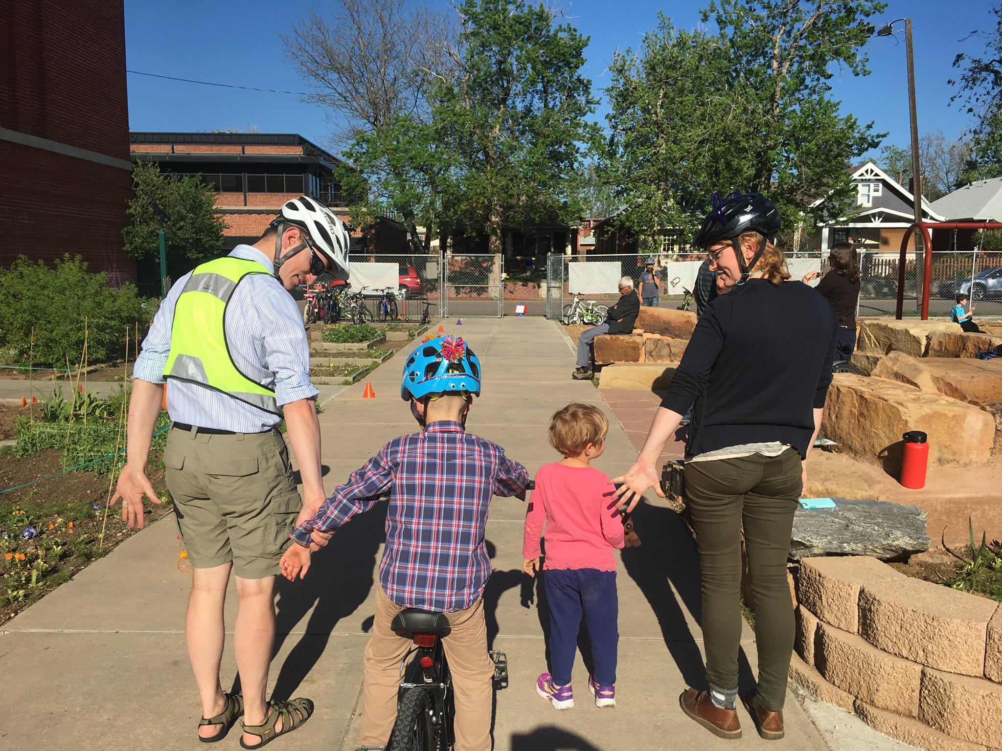 An adult wearing a yellow vest, a child wearing a checkered shirt, a smaller child with a pink shirt and another adult in a black sweatshirt stand facing away from the camera. They are on a sidewalk.