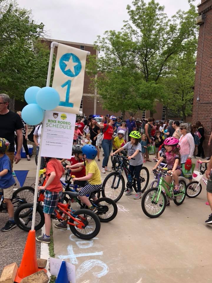 A number of children with bikes and helmets stand over their bikes on paved ground. There is a cone holding a sign and balloons in the foreground.