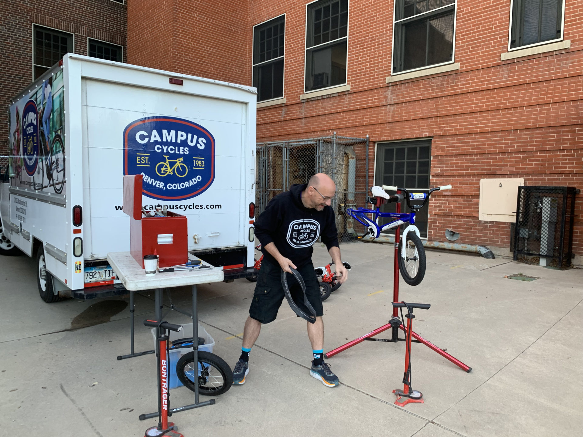 A man stands behind a vertical bike repair stand. There is a van behind him that has a logo for Campus Cycles. They are next to a red brick building.