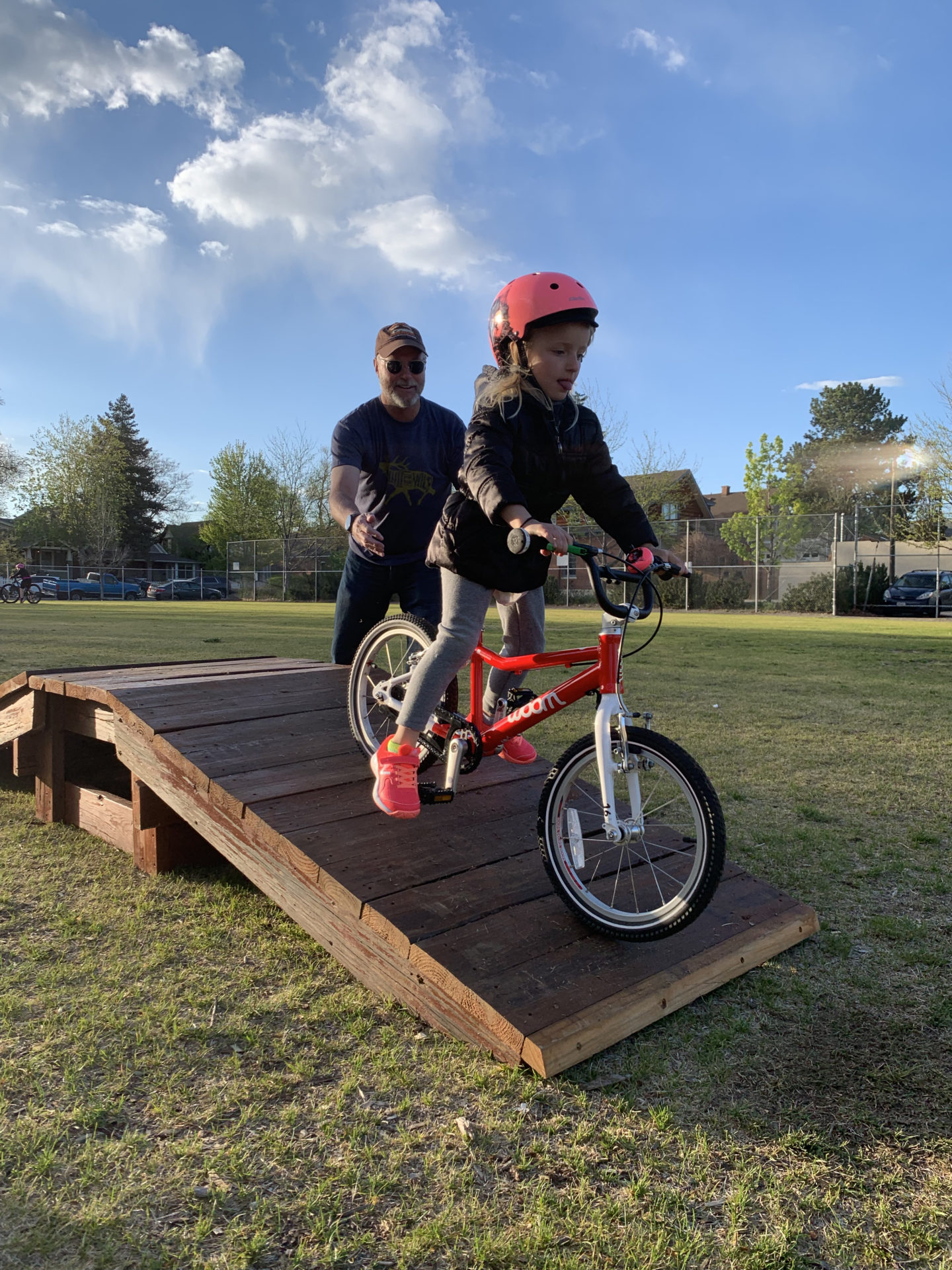 A child wearing a helmet rides down a wooden ramp over a grass field. There is an adult behind her.