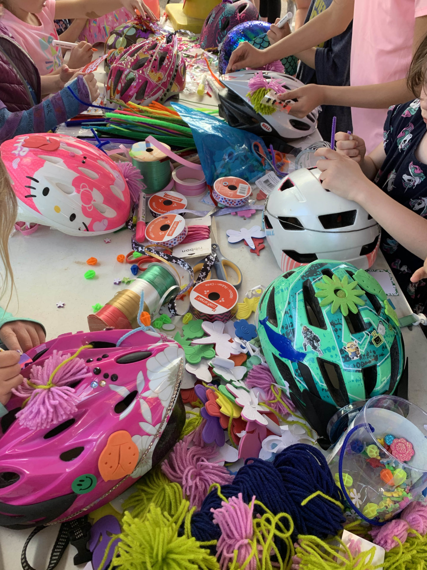 A number of helmets on a table. Children's hands are shown decorating the helmets with streamers, glitter, glue and other materials.