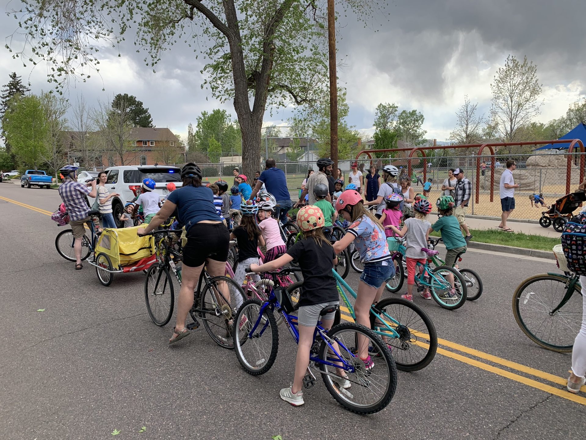 A group of adults and children riding bikes away from the camera toward the left, on a road. The school playground is in the background.