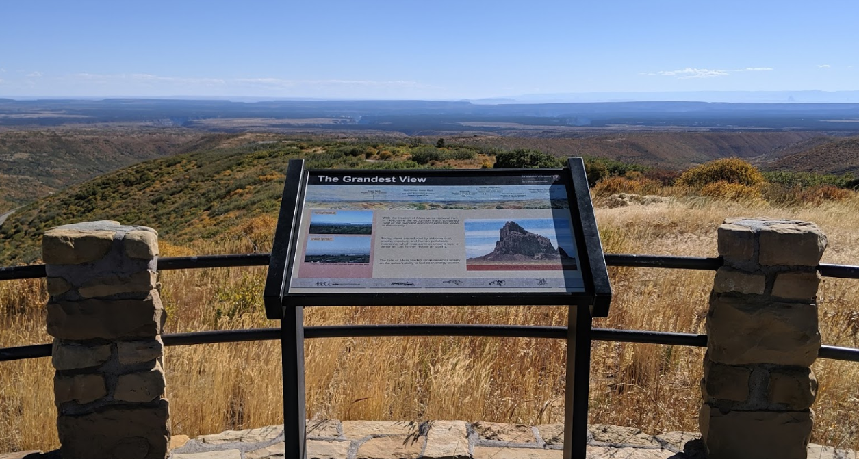An overlook point looking over hills and distant cliffs. There is a sign with information about the view in the foreground but the text is not legible in this photo.