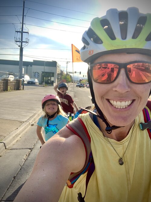 A person taking a selfie. They are wearing a helmet, sunglasses, a tank top and they are smiling. Behind them are two children also wearing helmets and smiling. They are all on the shoulder of a road, next to a sidewalk.