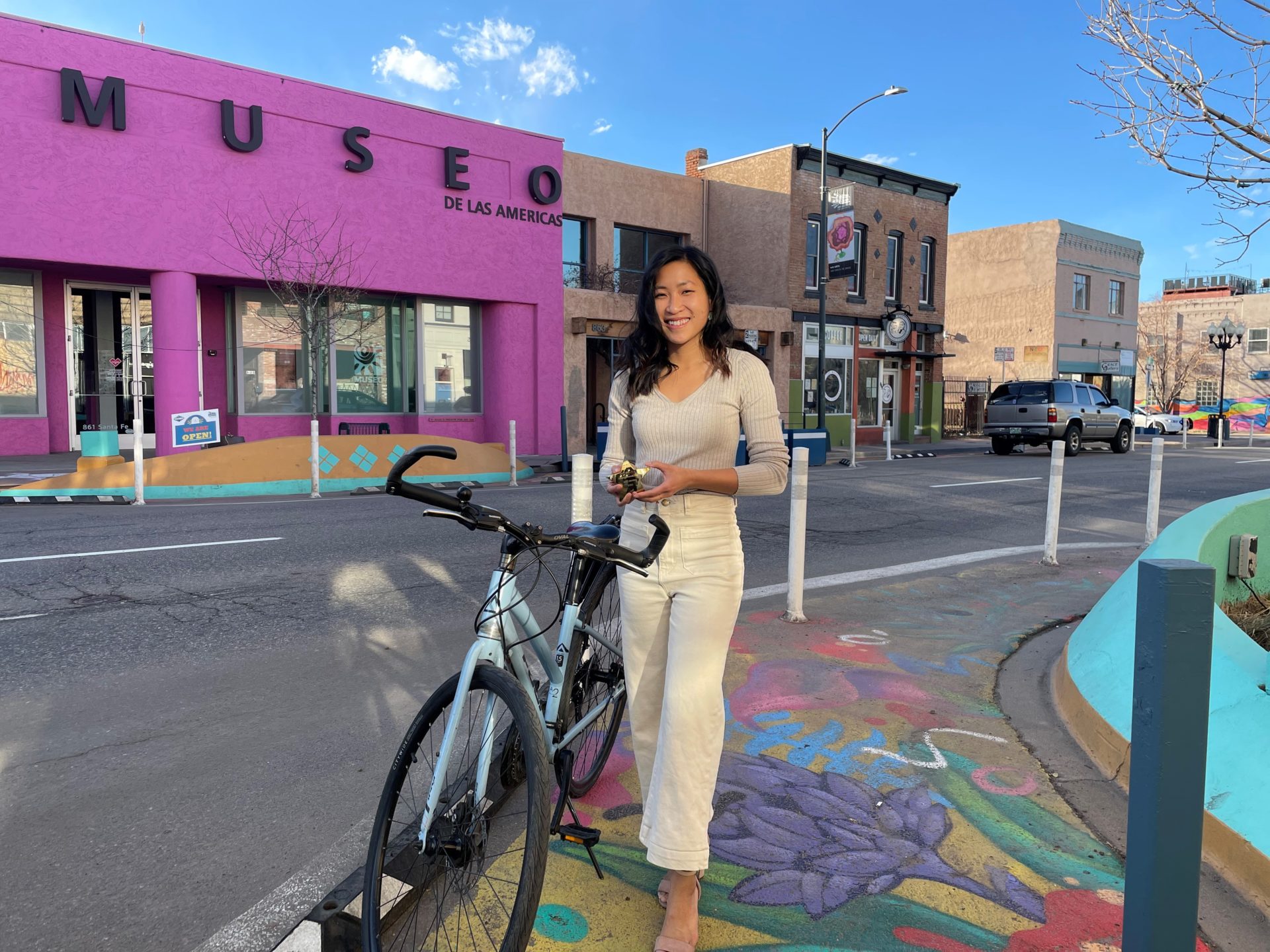 A woman stands with her bike next to her in front of a street, with a bright pink building in the background.