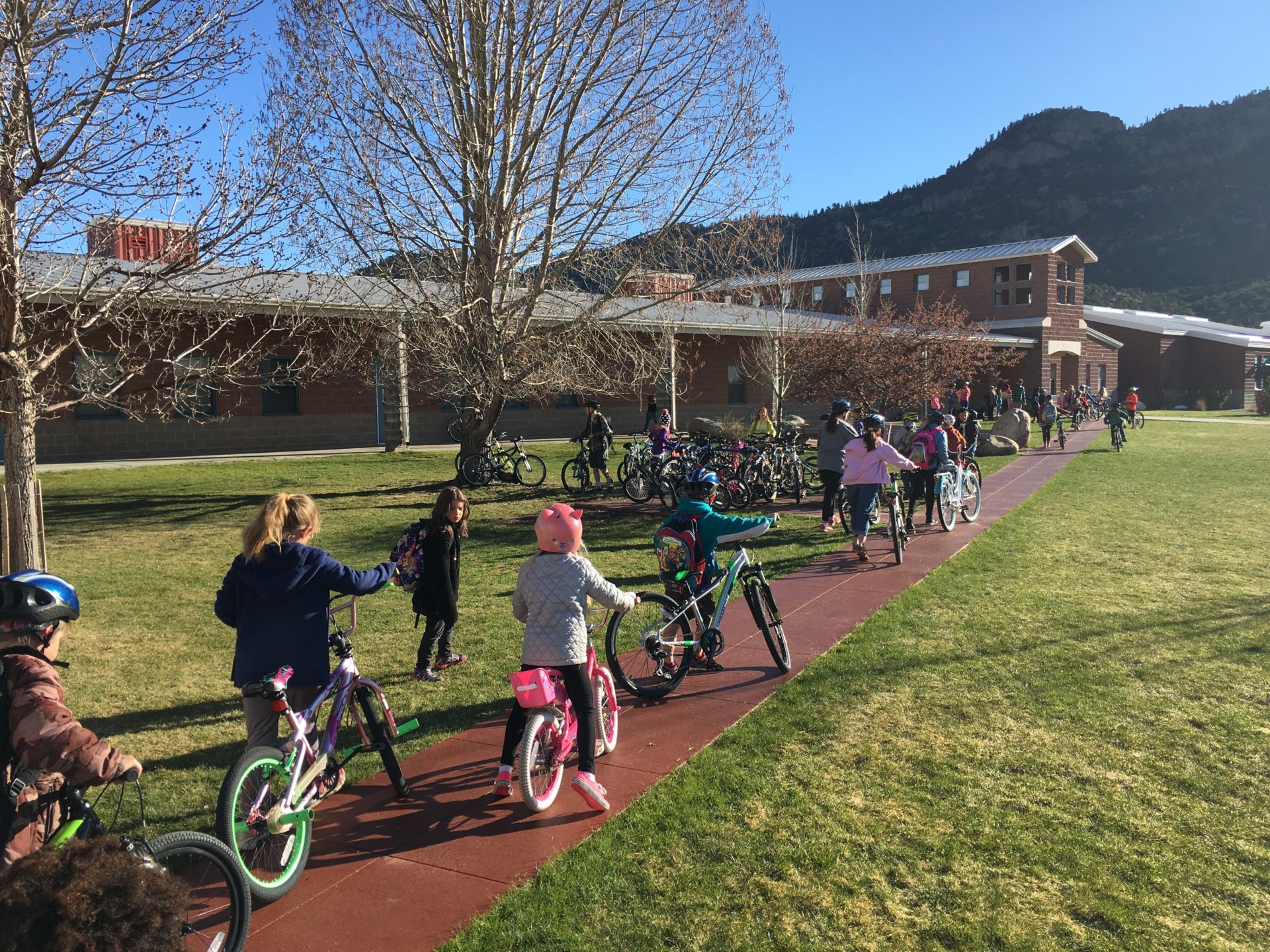 A group of children ride bikes up a sidewalk toward a school building in the background.