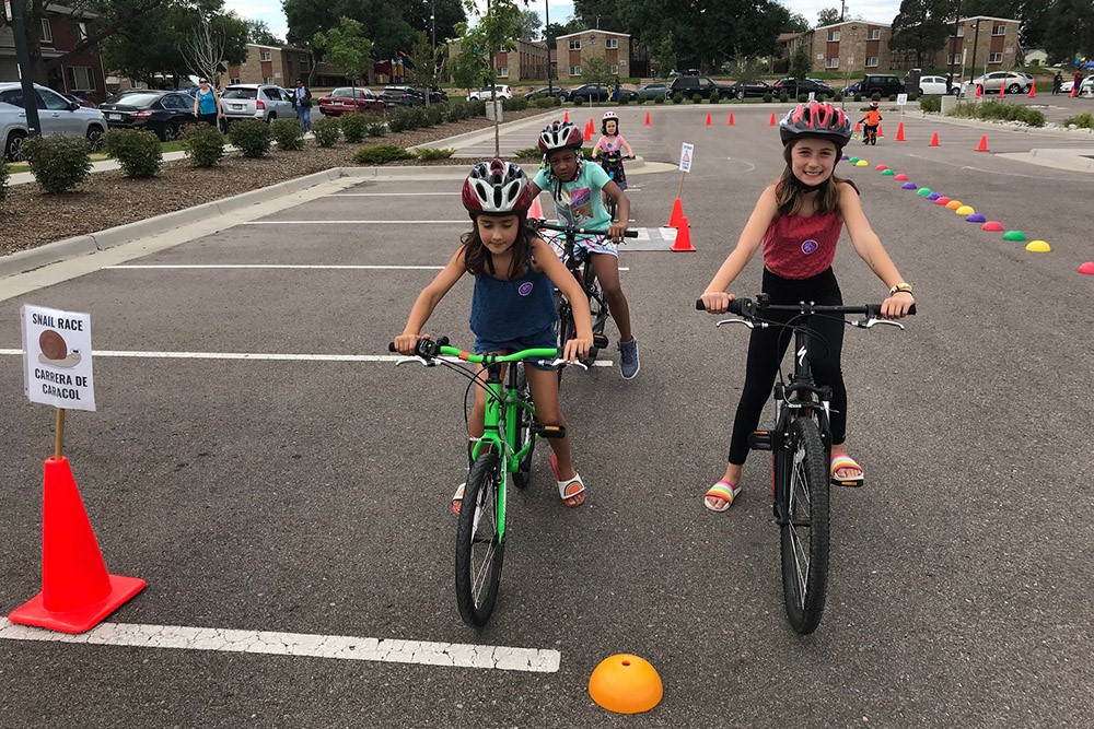 A few children ride bikes toward the camera in a parking lot. There are some cones and small domes on the ground nearby.