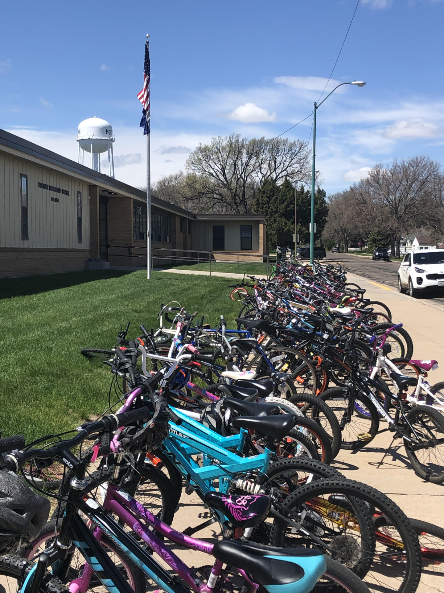 A row of bicycles parked on pavement in the center of the image. On the left side is a lawn and then a building.