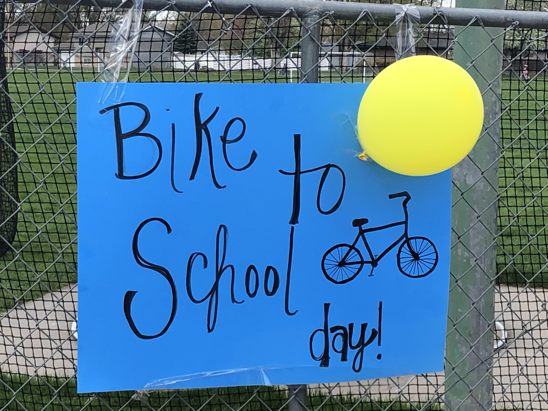 A sign on blue paper mounted on a fence, with a yellow balloon attached on the top right of it. It reads "Bike to School Day!" and there's a drawing of a bicycle.