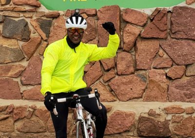 A person in a yellow cycling jacket on a bike has a hand raised in a fist. They are in front of the Pikes Peak Summit sign.