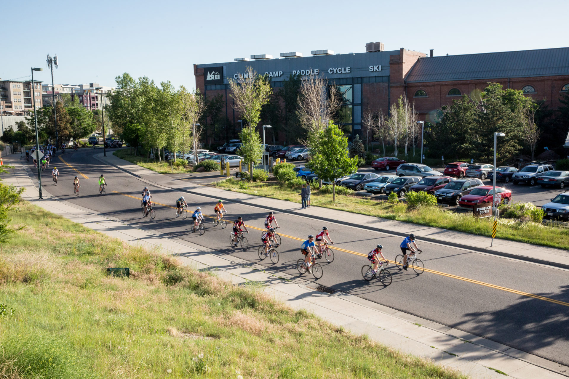 A group of people ride bikes on a paved road. There is an REI store in the background.