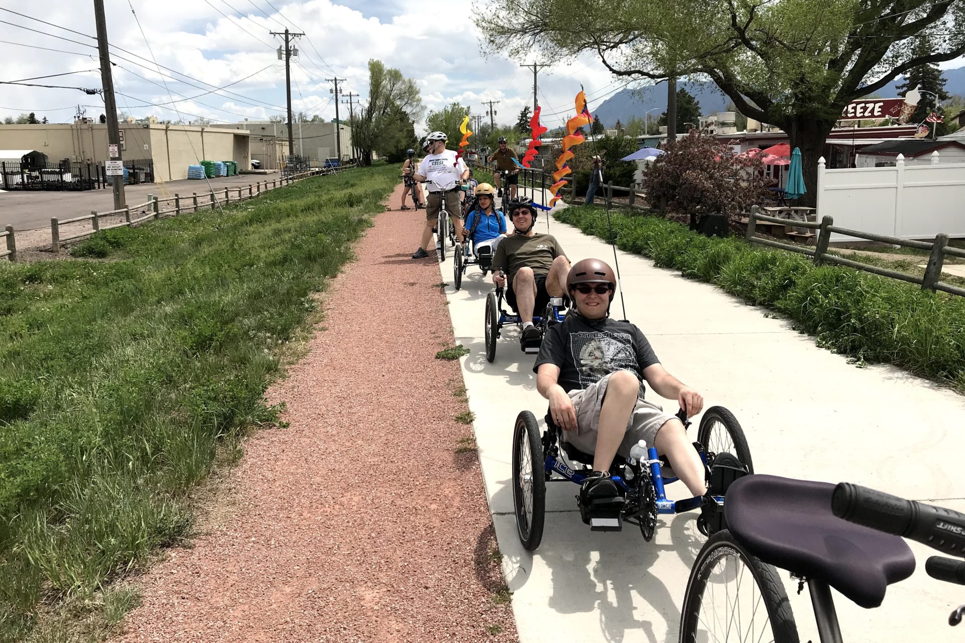 A group of people parked on adaptive trikes in a line front to back on a paved trail.