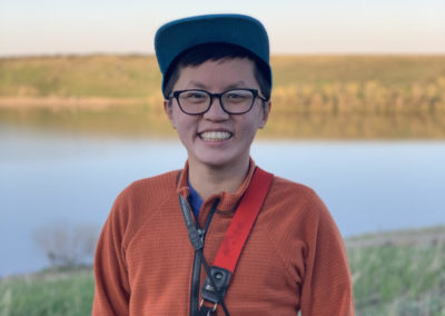 A photo of a person from the legs up. They are wearing a baseball cap and prescription glasses and are smiling with their teeth. They have a camera strapped around their neck.