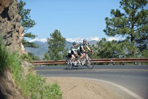 Two people ride a tandem bike on a paved mountain road.
