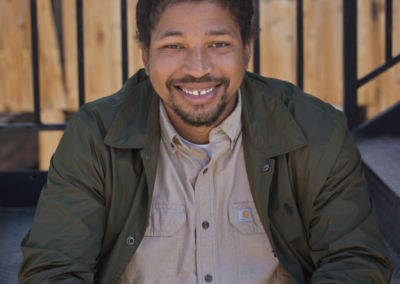 A person photographed from the waist up smiling at the camera. They are sitting on a stoop. They are smiling with their teeth.
