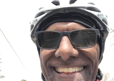 A person smiling with their teeth at the camera, photographed from the chin up. They are wearing a helmet and sunglasses.