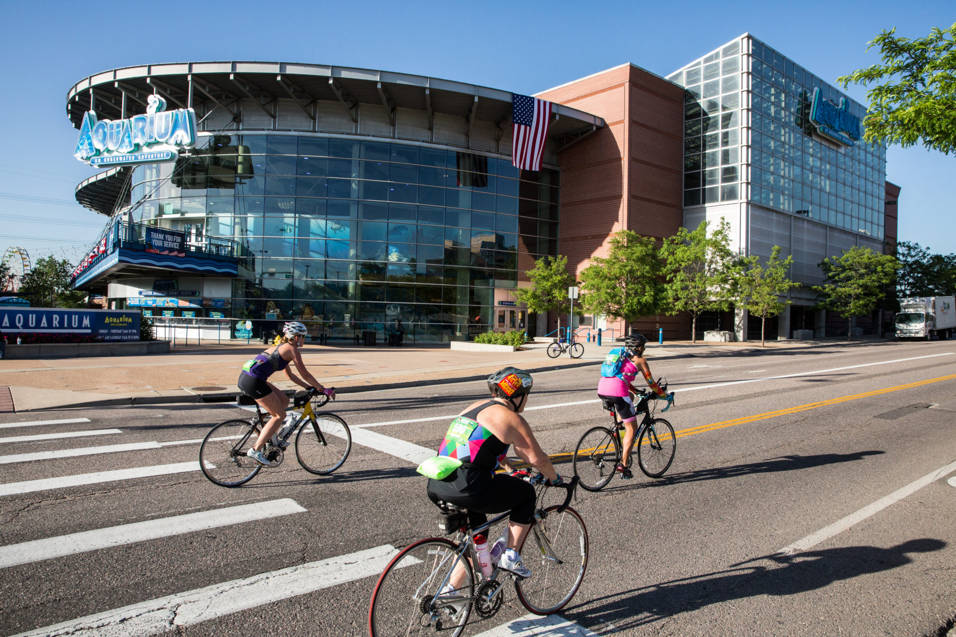 Three people ride bikes on a road toward the right of the frame. The Denver Aquarium is behind them.