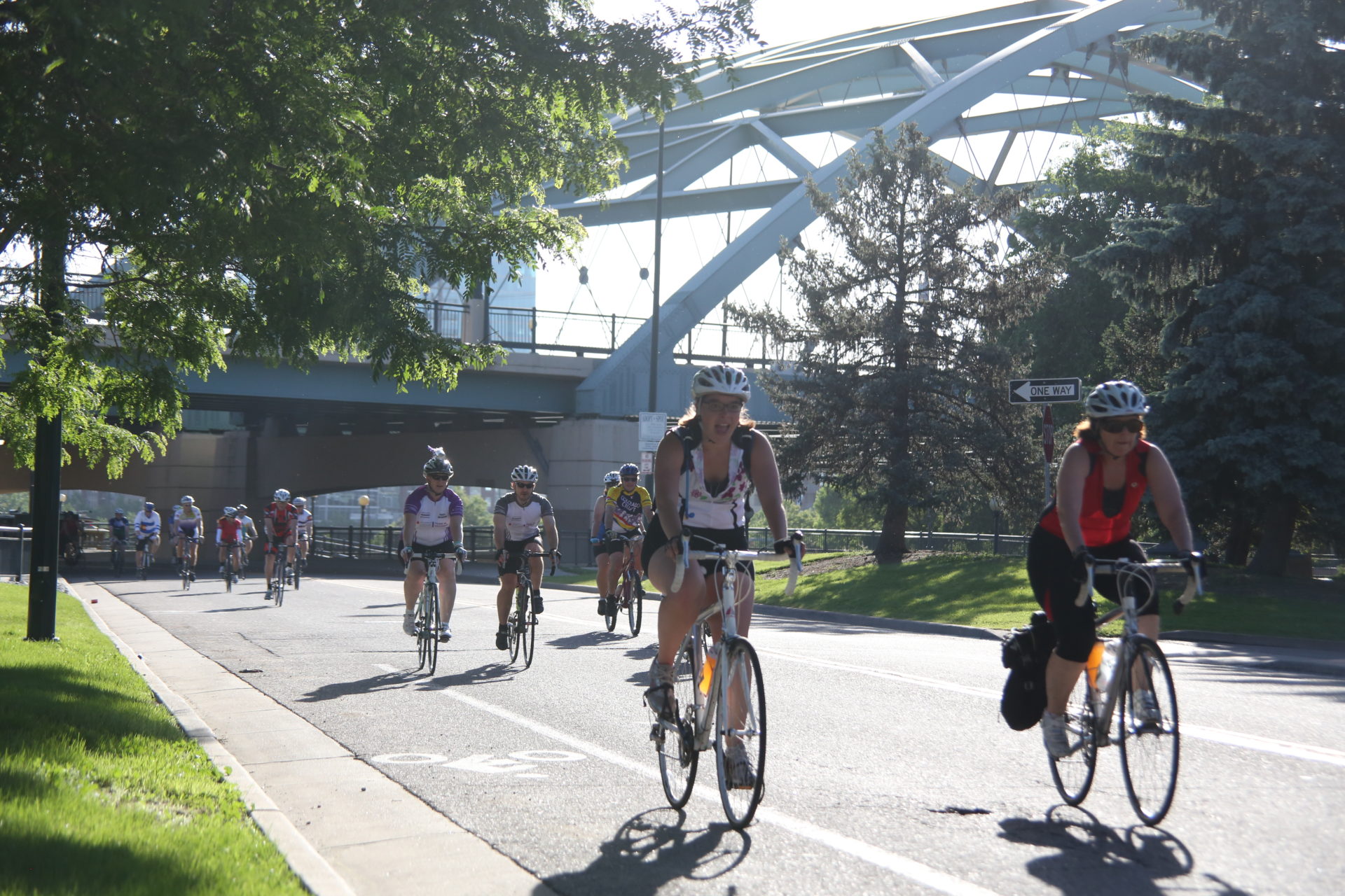 A group of people ride bikes on a paved road to the foreground right of the frame. In the background, there is a bridge under which the riders are biking. 