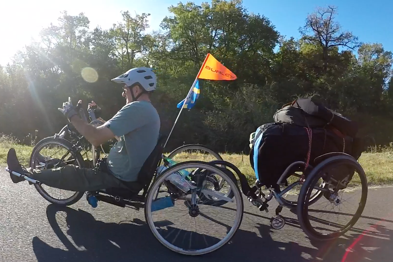 A person riding a handcycle rides toward the left of the frame. They are towing their wheelchair with bags on it behind them.